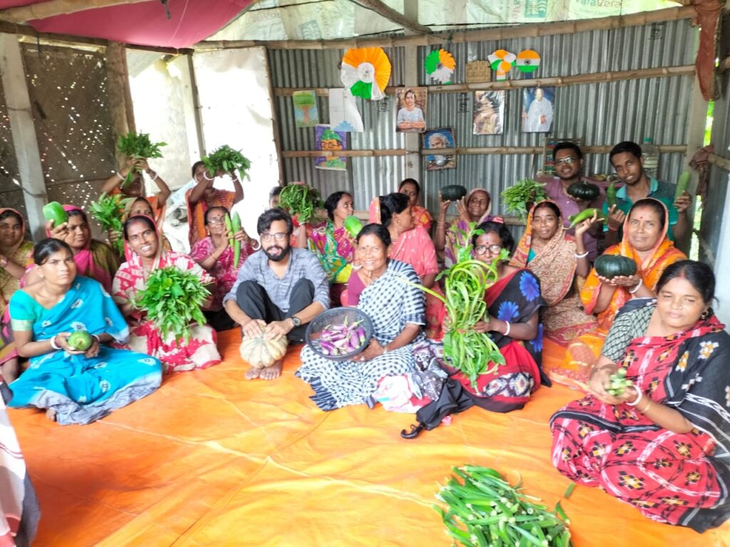 Sitting with a group of women while holding vegetables grown by them